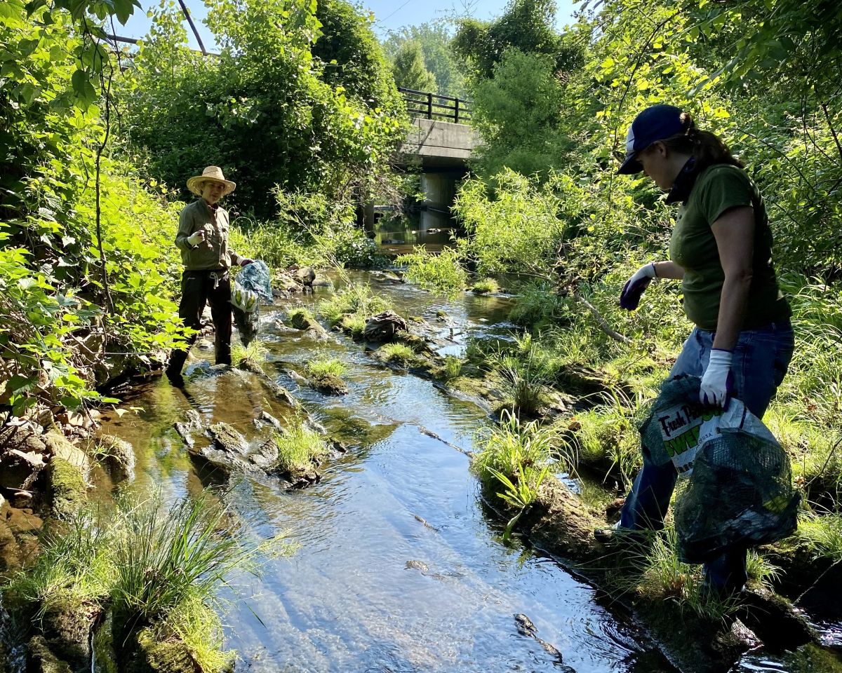 two people walk through a stream holding bags of trash they've collected